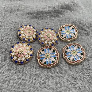 May include: Six decorative buttons with floral designs. The buttons have gold-coloured metal rims, with white, pink, blue, and green enamel details. The buttons are round and have a vintage aesthetic.