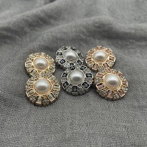 May include: Six decorative buttons with a pearl center and a surrounding design of small, sparkling rhinestones. The buttons are in gold, silver, and rose gold tones, arranged on a gray fabric background.