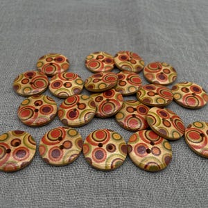 Puede incluir: Una colección de botones de madera con un diseño circular en tonos rojos, naranjas, verdes y morados. Cada botón tiene dos agujeros para coser. Los botones están dispersos sobre una tela gris.