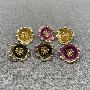 以下が含まれることがあります： 金、紫、黒色の花型のスタッドピアス6個。 イヤリングは、小さな丸い中心部を備えた、テクスチャ加工された、層状の花びらのデザインです。 イヤリングは、グレーのテクスチャ加工された表面に配置されています。