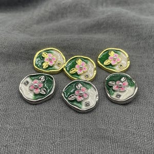 May include: Six decorative floral buttons with a pearl centre and pink petals. The buttons have a white and green enamel design with gold or silver metal edging. The buttons are arranged on a grey fabric surface.