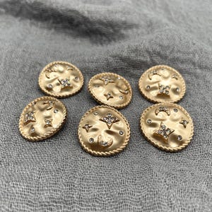 May include: Six gold-coloured buttons with celestial designs. Each button features a textured surface with a star, moon, and crystal accents. The buttons have a rope-like edge and are arranged on a grey fabric.