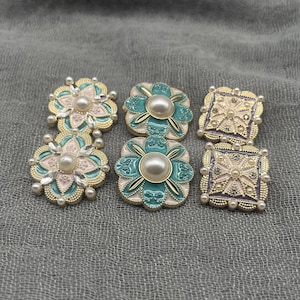 May include: Six decorative buttons featuring gold-tone metal edges and faux pearl centers. Two buttons are flower-shaped with pink and teal enamel, two are teal floral shapes, and two are square with pink and purple enamel accents.