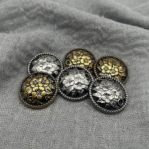 Pode incluir: Um conjunto de seis botões redondos de metal com desenhos florais. Os botões apresentam tons prateados e dourados, com detalhes intrincados nas pétalas e uma borda decorativa. Estão dispostos sobre um fundo de tecido cinzento.