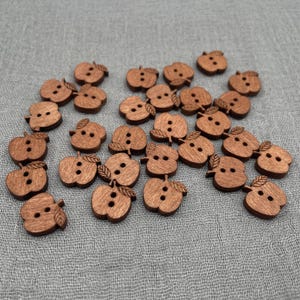 Könnte beinhalten: Eine Sammlung von holzfarbenen Knöpfen in Apfelform. Jeder Knopf hat eine warme braune Farbe, zwei oder drei Löcher zum Annähen und ein kleines Blattdetail. Die Knöpfe sind auf einem grauen Stoff verteilt.
