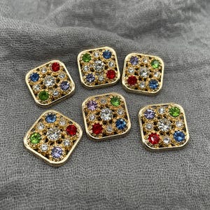 May include: Six square gold-toned buttons with a central clear crystal surrounded by smaller, colorful gemstones. The gemstones are red, green, blue, and purple. The buttons are arranged on a gray fabric.