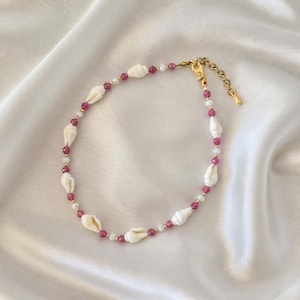 Op de afbeelding: Een kralenketting met witte schelpen afgewisseld met kleine, gefacetteerde magentakleurige kralen, piepkleine witte kraaltjes en goudkleurige metalen tussenstukjes. De ketting heeft een goudkleurige karabijnsluiting en een verstelbare kettingverlenging.