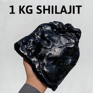 May include: A dark, glossy black mass of Shilajit, held in a hand against a white background. The text "1 KG SHILAJIT" is printed in bold black letters above the object. The substance appears to be a solid, natural resin.