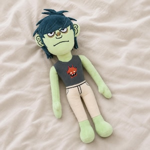 May include: A plush doll of a cartoon character with green skin, blue hair, and a stern expression. The doll wears a gray tank top with a red and orange graphic and cream-colored pants. The doll is lying on a cream-colored surface.