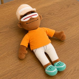 May include: A plush doll of a person wearing an orange shirt, white pants, and teal shoes. The doll has a tan hat and sunglasses. The doll is lying on a wooden surface.