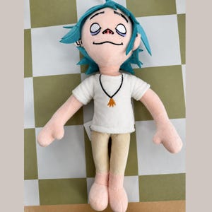 May include: A plush doll of a cartoon character with blue spiky hair, white t-shirt, tan pants, and pink limbs. The doll has a black necklace with a gold pendant. The doll's face has black facial features and closed eyes.