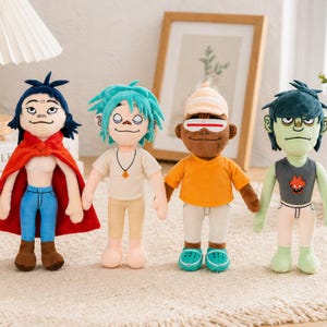 May include: Four plush dolls of cartoon characters. Each doll has a unique hair color and outfit. One doll wears a red cape and blue jeans. Another has turquoise hair and a white shirt. The third doll has orange clothing and the fourth has green skin.