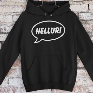Könnte beinhalten: Schwarzer Kapuzenpullover mit einer weißen Sprechblase mit der Aufschrift "HELLUR!". Der Pullover hat eine Vordertasche und eine Kapuze mit Kordelzug. Der Hintergrund ist eine Backsteinmauer.