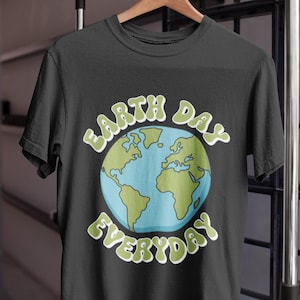 May include: Black t-shirt with the words "Earth Day Everyday" in green lettering. The design features a blue and green globe in the center. The shirt is hanging on a wooden hanger.