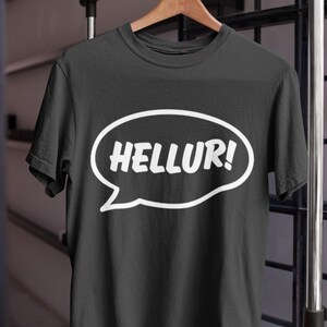 Könnte beinhalten: Schwarzes T-Shirt mit dem Wort "HELLUR!" in einer weißen Sprechblase. Das Shirt hängt an einem Holzbügel. Der Hintergrund umfasst eine Metallstange und einen dunklen Fensterrahmen.