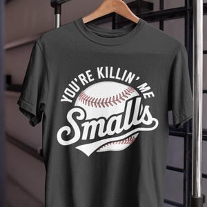 Op de afbeelding: Zwart t-shirt met een honkbalafbeelding. Het ontwerp toont een honkbal met de woorden "You're Killin' Me Smalls" in witte letters. Het shirt hangt aan een houten hanger.