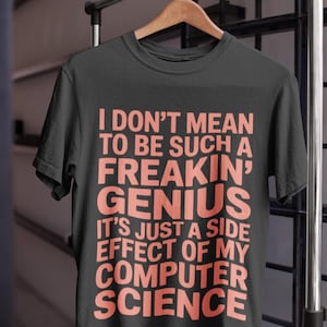 Può includere: T-shirt nero con la scritta "I DON'T MEAN TO BE SUCH A FREAKIN' GENIUS IT'S JUST A SIDE EFFECT OF MY COMPUTER SCIENCE" in lettere color corallo. La maglietta è appesa a una gruccia di legno.