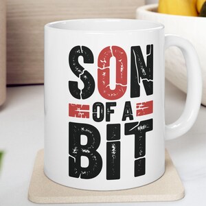 May include: A white ceramic mug featuring the text "SON OF A BIT" printed in a bold, distressed font. The word "SON" and "BIT" are in black, while the "O" and the horizontal bars flanking "OF A" are in a textured, weathered red finish.