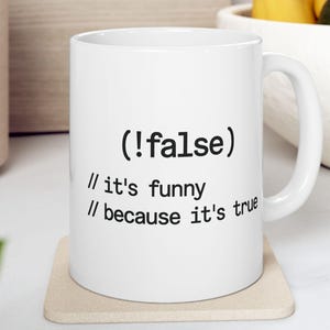 Op de afbeelding: Een witte keramische mok met zwarte bedrukte tekst die (!false) leest, gevolgd door twee regels codecommentaar met de tekst // it's funny en // because it's true. De mok heeft een standaard cilindrische vorm met een gebogen handvat.