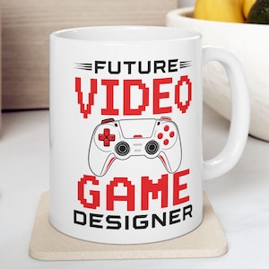 May include: A white ceramic mug featuring a graphic of a video game controller in the center. The text "FUTURE" appears above the controller, and "VIDEO GAME DESIGNER" is printed below it in a bold, red, pixelated font style.