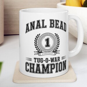 Può includere: Una tazza in ceramica bianca con testo nero che recita ANAL BEAD 1 TUG-O-WAR CHAMPION. Il design include un emblema circolare centrale con il numero 1 circondato da una corona d'alloro, il tutto in un carattere audace in stile college.