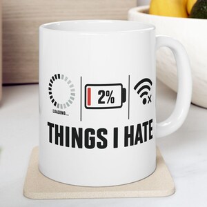 May include: A white ceramic mug featuring a black graphic design with a loading icon, a battery icon at 2% charge, and a disconnected Wi-Fi symbol. Below these icons, the text reads THINGS I HATE in bold, black, capital letters on the front.