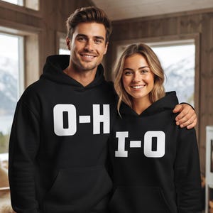 May include: Two black hooded sweatshirts featuring white block lettering. The left sweatshirt displays the text O-H, while the right sweatshirt displays the text I-O. Both garments include a front kangaroo pocket and long sleeves with ribbed cuffs.