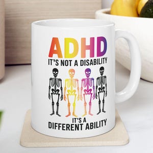 Peut inclure: Mug en céramique blanche avec le texte "ADHD IT'S NOT A DISABILITY IT'S A DIFFERENT ABILITY" en couleurs arc-en-ciel. La tasse présente quatre squelettes colorés. La tasse est posée sur un dessous de verre beige.