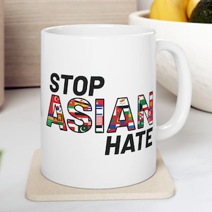 May include: White ceramic mug with the words "STOP ASIAN HATE" in black text. The word "ASIAN" is filled with various country flags. The mug is on a beige coaster.