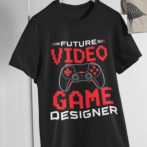Op de afbeelding: Een zwart T-shirt met korte mouwen en een rode pixelafbeelding van een videogamecontroller. De tekst FUTURE VIDEO GAME DESIGNER staat in een vet, blokkerig lettertype. Het ontwerp is gecentreerd op de borst van het T-shirt op een hanger.