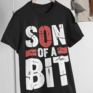 May include: A black short-sleeved t-shirt featuring the white and red distressed text "SON OF A BIT" printed on the front. The shirt is displayed on a wooden hanger, showcasing a crew neckline and a standard fit with a graphic design.