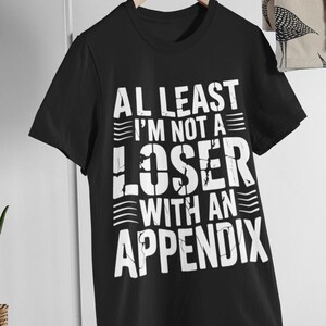 May include: A black short-sleeved t-shirt featuring white distressed-style text that reads: AT LEAST I'M NOT A LOSER WITH AN APPENDIX. The shirt is displayed on a wooden hanger against a plain background, showcasing a bold graphic design.