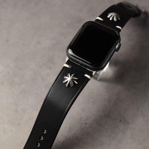 以下が含まれることがあります： 黒いApple Watch。黒い革製のバンドには、銀色の星型の装飾が施されています。バンドは白いステッチとクラシックなバックルが付いています。時計の文字盤は黒で、全体的にモダンでスタイリッシュなデザインです。