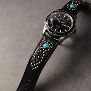 Puede incluir: Un reloj negro con una correa de cuero negro adornada con piedras de color turquesa y tachuelas plateadas. La esfera del reloj es negra con marcas blancas y la marca "Hamilton". La correa tiene un patrón floral.