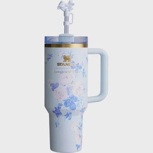 May include: A light blue insulated tumbler with a handle, adorned with a floral pattern in blue and pink hues. It includes a clear lid, a straw, and a decorative bow. The text "STANLEY" and "LOVESHACKF" are printed on the tumbler. The tumbler is designed for cold beverages.