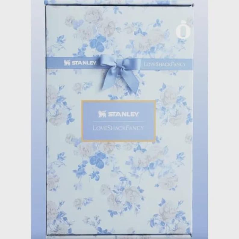 May include: A rectangular box with a floral pattern in shades of blue and white. The box features the "STANLEY" and "LOVESHACKFANCY" logos. A light blue ribbon with a bow is tied around the box, adding a decorative touch.