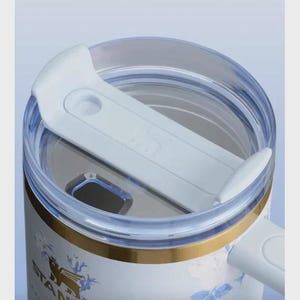 May include: Close-up of a white and blue floral Stanley tumbler with a clear lid and a white slider. The tumbler has a gold band and a handle. The Stanley logo is visible on the side.