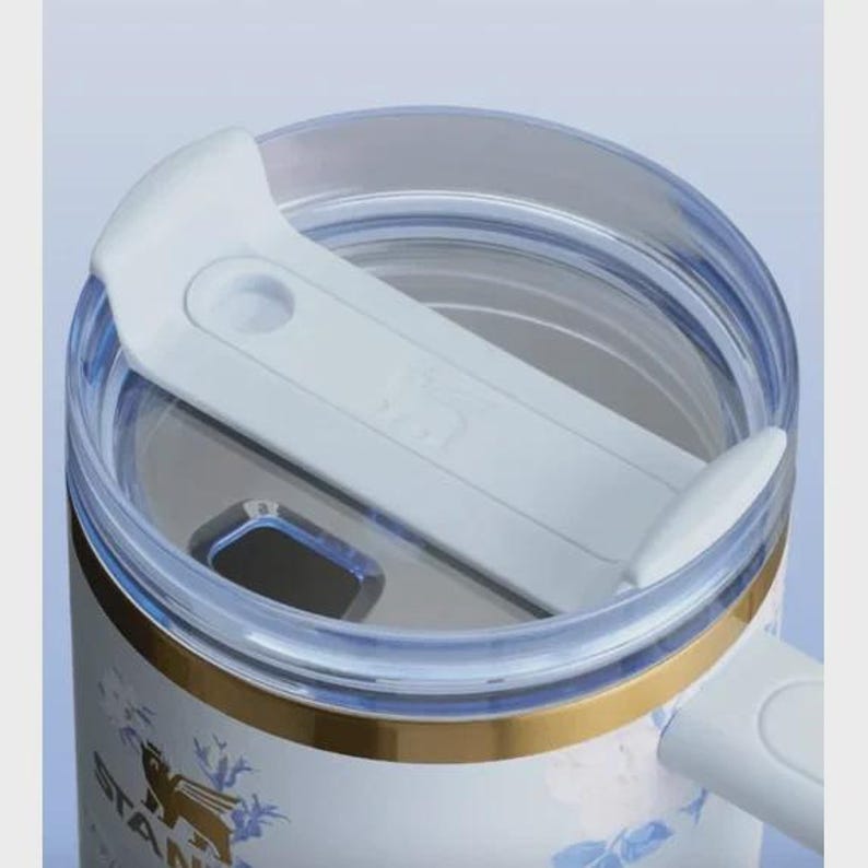 May include: Close-up of a white and blue floral Stanley tumbler with a clear lid and a white slider. The tumbler has a gold band and a handle. The Stanley logo is visible on the side.