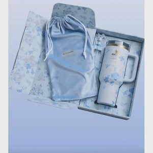 May include: A white insulated travel mug with a handle, decorated with blue floral designs, presented in a floral-patterned box. Includes a matching blue drawstring bag. The mug has the brand name "STANLEY" printed on it.