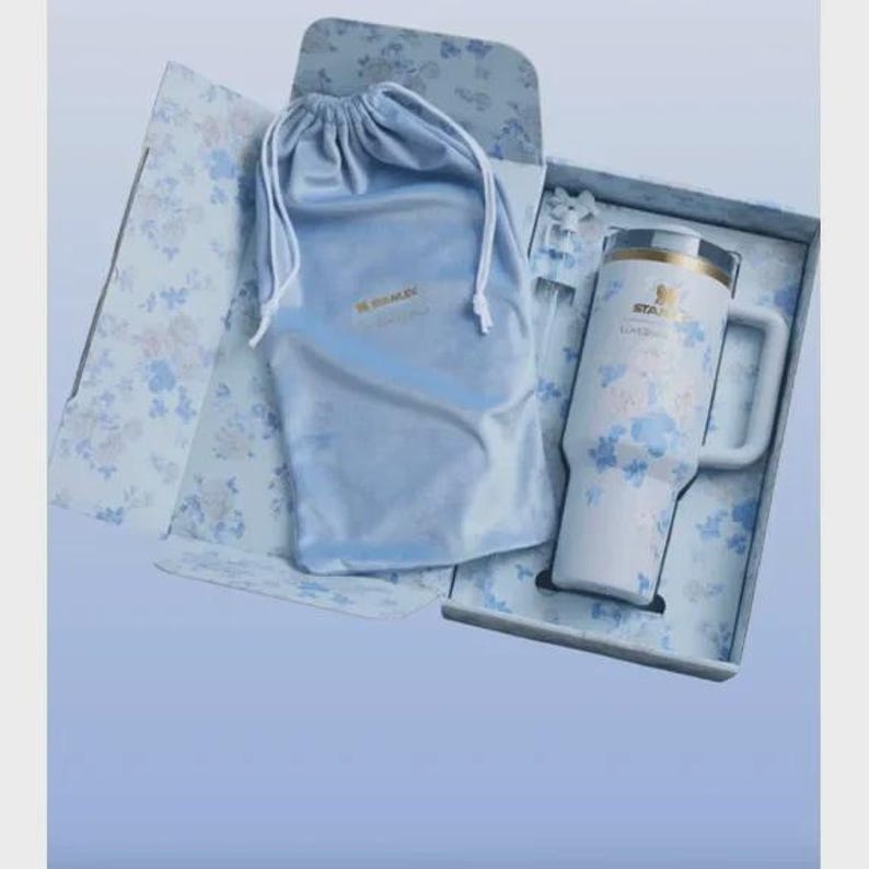 May include: A white insulated travel mug with a handle, decorated with blue floral designs, presented in a floral-patterned box. Includes a matching blue drawstring bag. The mug has the brand name "STANLEY" printed on it.