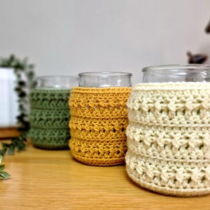 May include: Three glass jars with decorative crochet sleeves in olive green, mustard yellow, and cream. The crochet sleeves have a textured, openwork design. The jars are on a wooden surface, with greenery in the background.