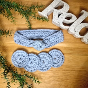 May include: A light blue crocheted headband with a knotted design and matching coasters. The word "Relax" is spelled out in large, textured letters. Green sprigs are in the corner. The items are on a wooden surface.