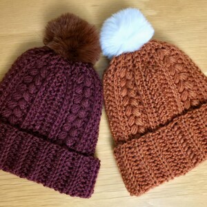 以下が含まれることがあります： 2つのニット冬用帽子。1つは、茶色のフェイクファーのポンポンが付いた濃いバーガンディ色です。もう1つは、白いフェイクファーのポンポンが付いた、焦げたオレンジ色です。どちらの帽子にも折り返しがあります。