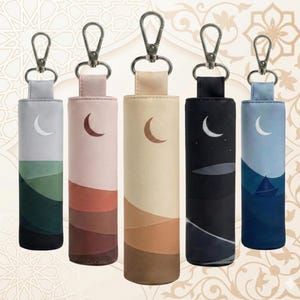 May include: Five cylindrical keychains with a landscape design. Each keychain features a different color scheme, including gray, pink, beige, black, and blue. Each keychain has a crescent moon design and a silver clasp.