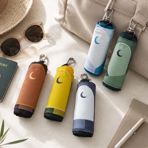 May include: A collection of compact, rolled-up umbrellas in various colors, including blue, green, yellow, and brown, each featuring a crescent moon design. The umbrellas are attached to silver carabiner clips. Other travel accessories, such as a passport, sunglasses, and a hat, are also visible.