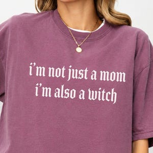 May include: A berry-coloured short-sleeve t-shirt featuring the white text "I'm not just a mom I'm also a witch" printed in a gothic-style font across the chest. The garment has a crew neckline and a relaxed fit, worn with a gold chain necklace.
