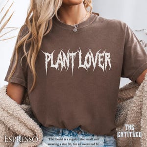 以下が含まれることがあります： エスプレッソ色のTシャツ。白のゴシック体フォントで「PLANT LOVER」と書かれています。「THE ENTITLED」コレクションの一部です。モデルはオーバーサイズフィットのためにXLサイズを着用しています。