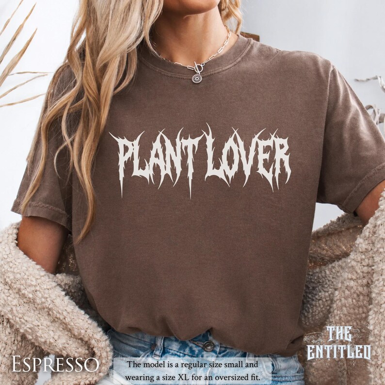以下が含まれることがあります： エスプレッソ色のTシャツ。白のゴシック体フォントで「PLANT LOVER」と書かれています。「THE ENTITLED」コレクションの一部です。モデルはオーバーサイズフィットのためにXLサイズを着用しています。