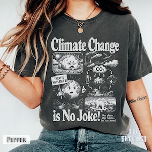 May include: A charcoal gray t-shirt featuring a graphic print with the text Climate Change is No Joke. The design includes illustrations of a sweating earth, a polar bear, CO2 emissions, and a sign that reads There's No Planet B.