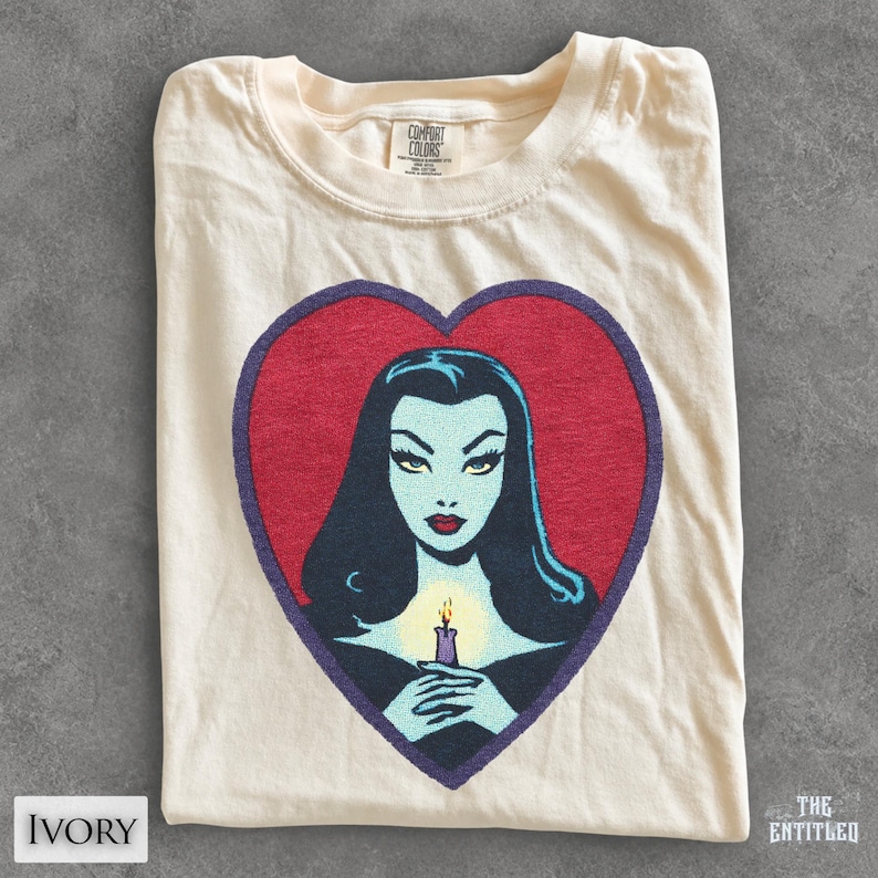 May include: An ivory t-shirt featuring a large red heart graphic with a dark purple outline. Inside the heart is a stylized illustration of a pale-skinned figure with long black hair, red lips, and a lit candle held in their hands. The shirt has a Comfort Colors label.